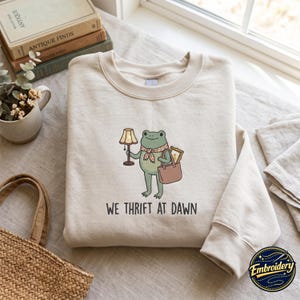 Puede incluir: Una sudadera beige de cuello redondo con un bordado de una rana verde con bufanda estampada, que sostiene una lámpara y un bolso. El texto "WE THRIFT AT DAWN" está bordado en color negro debajo de la ilustración de la rana en el frente.