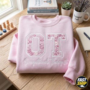 May include: A light pink sweatshirt with the embroidered letters "OT" in a floral design. The words "Occupational Therapist" are embroidered below the letters. The sweatshirt is on a wooden table, with books and a mug in the background.