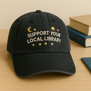 May include: Black baseball cap with the text "SUPPORT YOUR LOCAL LIBRARY" in gold lettering. The design includes a crescent moon, stars, and a sun. The cap is sitting on a wooden surface.