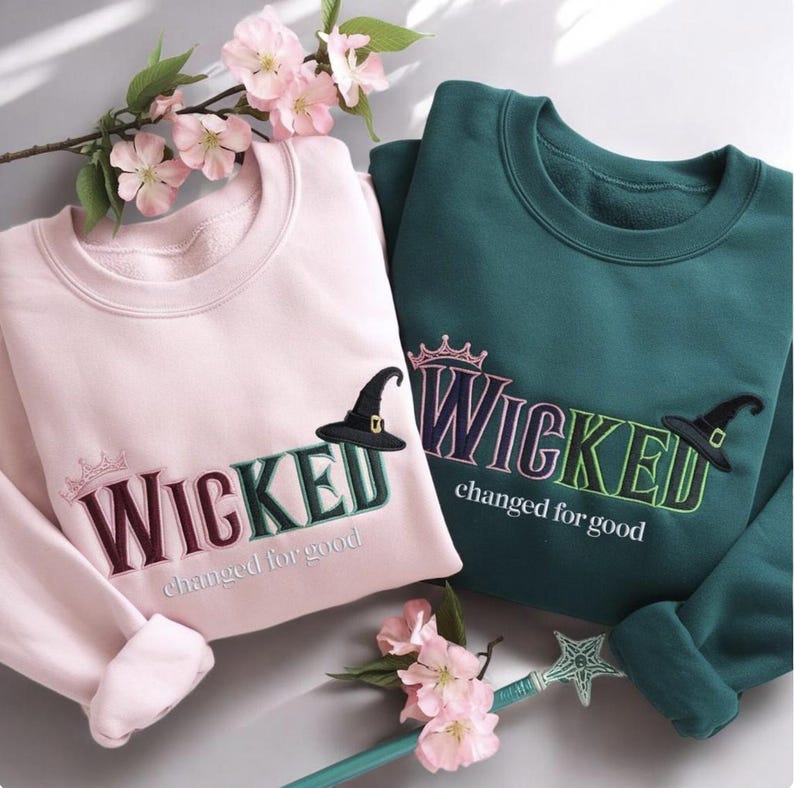 Puede incluir: Dos sudaderas de cuello redondo, una rosa claro y otra verde azulado, con la palabra "WICKED" bordada en la parte delantera. Cada sudadera presenta un sombrero de bruja y la frase "changed for good". Tambi&eacute;n hay flores decorativas y una varita.