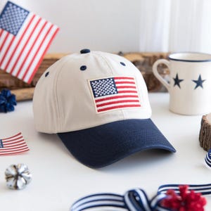 May include: A cream-coloured baseball cap with a navy blue brim features an embroidered American flag patch. The cap has navy blue eyelets and a button on top. Additional patriotic decorations, including flags and a mug with stars, are visible.