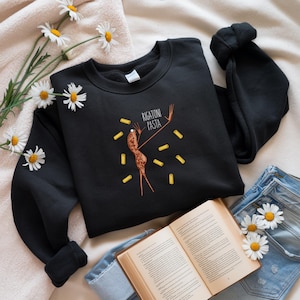 May include: Black sweatshirt featuring a whimsical illustration of a rigatoni pasta figure with outstretched arms, surrounded by scattered pasta shapes. The text "RIGATONI PASTA" is above the figure. The sweatshirt is displayed on a neutral surface with daisies and a book.