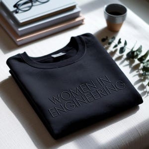 Könnte beinhalten: Ein schwarzes Sweatshirt mit dem gestickten Text "WOMEN IN ENGINEERING" liegt auf einer weißen Oberfläche. Das Rundhals-Sweatshirt steht im Mittelpunkt, mit Büchern, Brille und einer Kaffeetasse im Hintergrund.