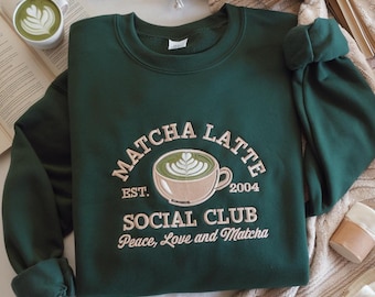 May include: A dark green sweatshirt with the words "Matcha Latte Social Club" embroidered in cream-colored thread. The design includes a coffee cup with a matcha latte design. The text "Peace, Love and Matcha" is also embroidered on the sweatshirt.