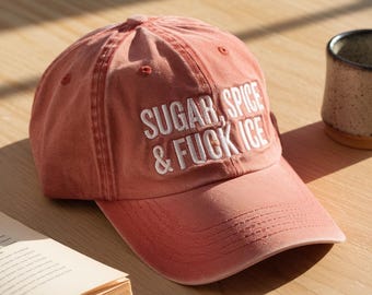 May include: A dusty rose baseball cap with the embroidered text "SUGAR, SPICE & FUCK ICE" in white. The cap is sitting on a wooden surface next to a book and a mug.