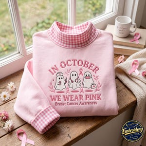 Op de afbeelding: Een lichtroze sweatshirt met een opstaande kraag en boorden in een roze-wit vichy-ruitpatroon. De voorzijde is voorzien van borduurwerk met de tekst IN OCTOBER WE WEAR PINK Breast Cancer Awareness en drie witte spoken met medische uitrusting.