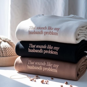 May include: Three folded sweatshirts in cream, black, and brown, stacked on top of each other. Each sweatshirt features the embroidered text "That sounds like my husband's problem" in a matching colour. The sweatshirts are neatly arranged.