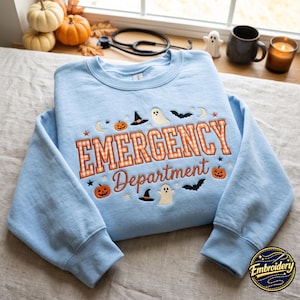 May include: A light blue crewneck sweatshirt featuring embroidered Halloween-themed designs including pumpkins, ghosts, bats, and witch hats. The text "EMERGENCY Department" is stitched in orange and white checkered and solid orange thread.