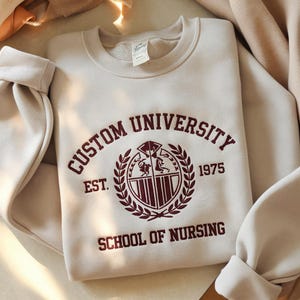 Könnte beinhalten: Ein beiger Pullover mit einem dunkelroten gestickten Logo, auf dem "Custom University Est. 1975 School of Nursing" steht.