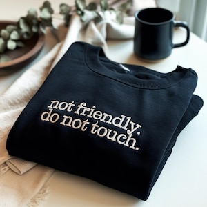 May include: Black sweatshirt with the embroidered text "not friendly. do not touch." A black mug and a wooden bowl with greenery are in the background. The sweatshirt is folded neatly on a white surface.