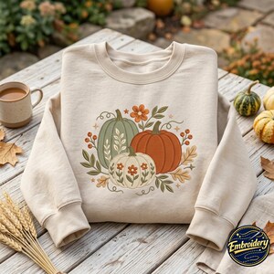May include: A beige crewneck sweatshirt featuring a central embroidered graphic of three pumpkins in sage green, orange, and cream. The design includes orange flowers, green leaves, and small berries, accented with delicate swirling vine details.