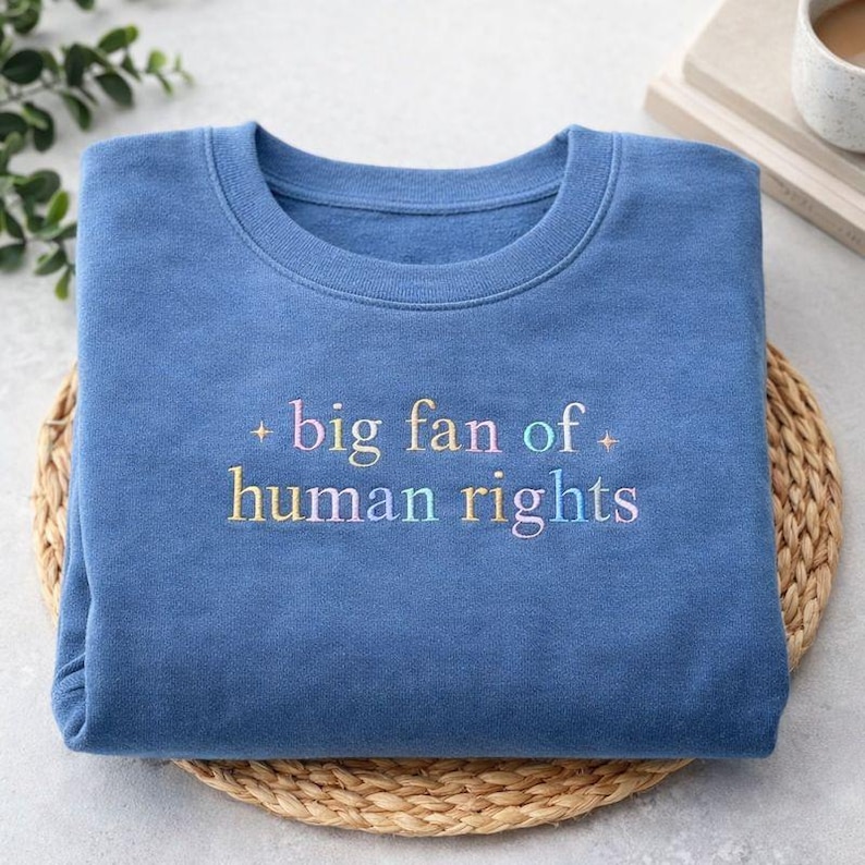 May include: A blue sweatshirt with the embroidered text "big fan of human rights" in pastel colors. The crew neck sweatshirt is folded neatly on a woven mat.