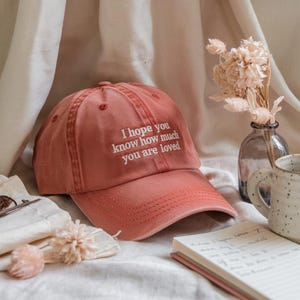 May include: A dusty rose baseball cap with the embroidered text "I hope you know how much you are loved." The cap is made of a soft fabric and has a curved brim. The hat is a casual accessory.