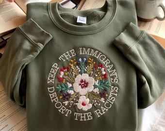 Może przedstawiać: Zielona bluza z haftowanym kwiatowym wzorem i napisem "KEEP THE IMMIGRANTS, DEPORT THE RACISTS". Haft przedstawia białe, czerwone i żółte kwiaty z zielonymi liśćmi. Bluza ma okrągły dekolt i długie rękawy.