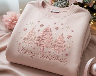 Puede incluir: Sudadera rosa claro con bordados de árboles de Navidad y copos de nieve. La frase "Merry Christmas" está bordada debajo. La sudadera está doblada sobre una superficie rosa, con decoraciones navideñas en el fondo.
