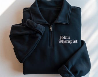 May include: A folded black sweatshirt with a quarter-zip collar. The words "Skin Therapist" are embroidered in white script on the chest. The sleeves are folded, and the sweatshirt is lying on a white surface.