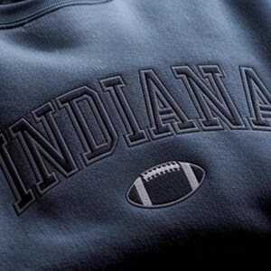 K&ouml;nnte beinhalten: Ein marineblaues Sweatshirt mit dem Wort "INDIANA" in einem dunklen, grauen Schriftzug im College-Stil &uuml;ber der Brust. Unter dem Text befindet sich eine Football-Grafik in Wei&szlig; und Grau.