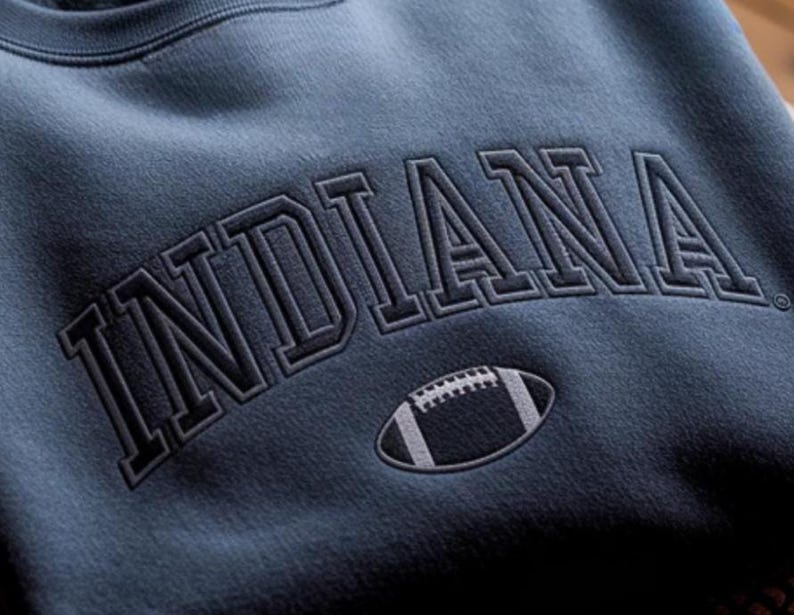 K&ouml;nnte beinhalten: Ein marineblaues Sweatshirt mit dem Wort "INDIANA" in einem dunklen, grauen Schriftzug im College-Stil &uuml;ber der Brust. Unter dem Text befindet sich eine Football-Grafik in Wei&szlig; und Grau.