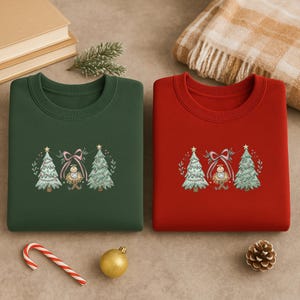 Puede incluir: Dos sudaderas de cuello redondo, una verde bosque y otra roja, cada una bordada con un diseño de árbol de Navidad. El diseño incluye dos árboles de Navidad, un lazo y una figura del niño Jesús. También se ven un bastón de caramelo, un adorno dorado y una piña.