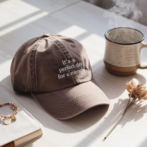Puede incluir: Gorra de béisbol marrón con el texto bordado "it's a perfect day for a miracle". La gorra está junto a una taza de café y una pulsera. También se ve una ramita de flores secas y un cuaderno.