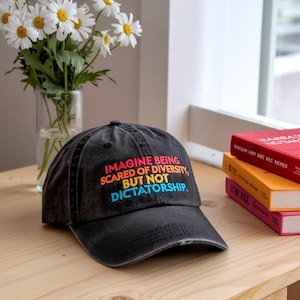 May include: A black baseball cap with a distressed look. The cap has a rainbow-colored embroidered message that reads "Imagine being scared of diversity, but not dictatorship."