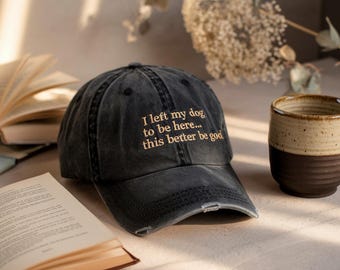 May include: A black distressed baseball cap with the text "I left my dog to be here... this better be good" embroidered in gold.