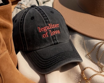 May include: A black baseball cap with the embroidered text "legalizer of love" in red. The cap is made of a worn-looking fabric. Other accessories, including a brown hat and jewelry, are in the background.