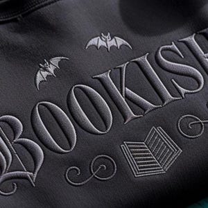 K&ouml;nnte beinhalten: Dunkelgraues Sweatshirt mit dem Wort "BOOKISH" in gotischer Schrift gestickt, begleitet von Fledermaus- und Buchillustrationen. Das Design ist in einem helleren Grau gehalten und bildet einen Kontrast zum dunklen Stoff. Ein gem&uuml;tliches und stilvolles Kleidungsst&uuml;ck f&uuml;r B&uuml;cherliebhaber.