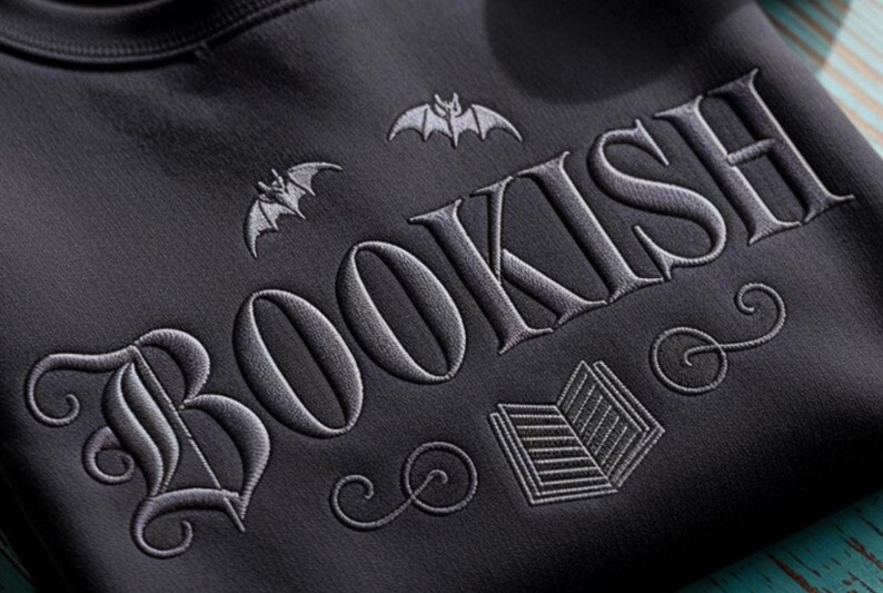 K&ouml;nnte beinhalten: Dunkelgraues Sweatshirt mit dem Wort "BOOKISH" in gotischer Schrift gestickt, begleitet von Fledermaus- und Buchillustrationen. Das Design ist in einem helleren Grau gehalten und bildet einen Kontrast zum dunklen Stoff. Ein gem&uuml;tliches und stilvolles Kleidungsst&uuml;ck f&uuml;r B&uuml;cherliebhaber.