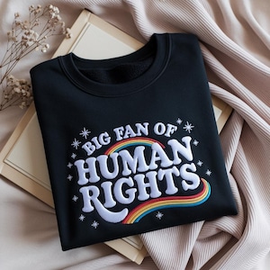 Op de afbeelding: Zwart sweatshirt met de geborduurde tekst "BIG FAN OF HUMAN RIGHTS" in het wit, met een regenboogontwerp. De sweatshirt heeft een ronde hals en wordt gepresenteerd op een neutrale achtergrond.