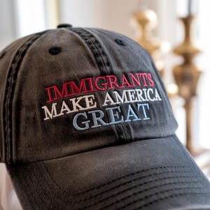 May include: A dark gray baseball cap with the embroidered text "IMMIGRANTS MAKE AMERICA GREAT" in red, white, and blue. The hat has a curved brim and a classic six-panel design.