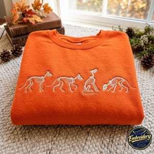 May include: A bright orange crewneck sweatshirt featuring white embroidered line art of four dog skeletons in various poses across the chest. The embroidery depicts the skeletal structures of dogs standing, sitting, and walking in a horizontal row.