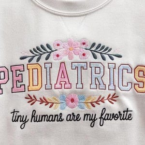 May include: White sweatshirt with embroidered text "PEDIATRICS" in rainbow colors and floral accents. The text "tiny humans are my favorite" is also embroidered below.