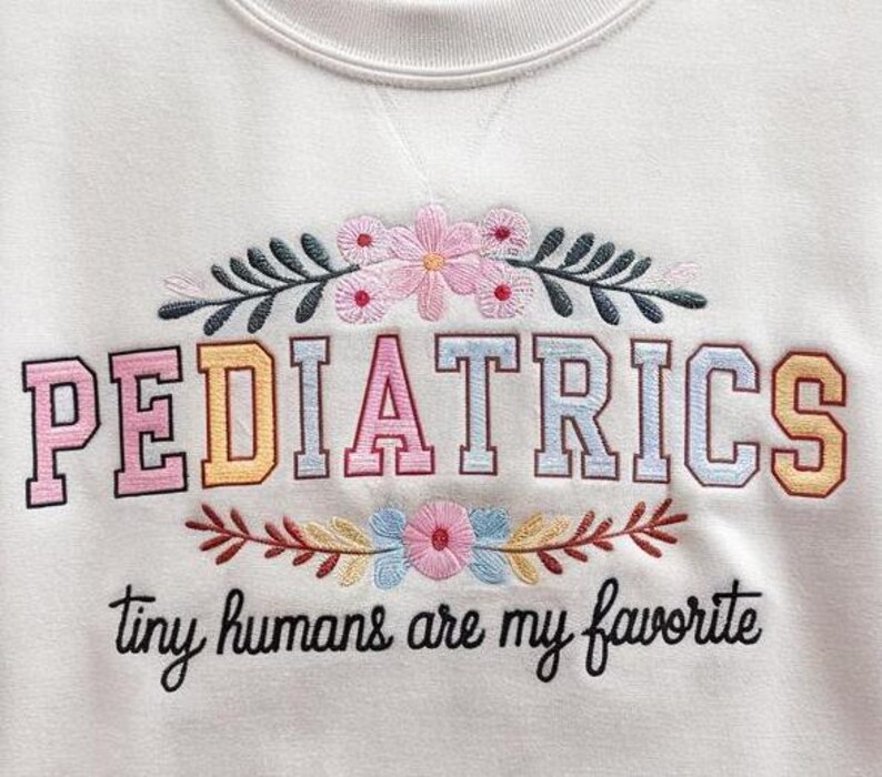May include: White sweatshirt with embroidered text "PEDIATRICS" in rainbow colors and floral accents. The text "tiny humans are my favorite" is also embroidered below.