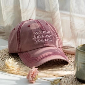 May include: A pink baseball cap with the text "women don't owe you shit" embroidered on the front.