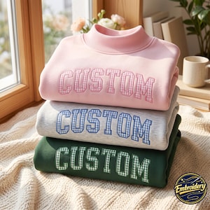 May include: A stack of three mock neck sweatshirts in light pink, heather gray, and dark green. Each sweatshirt features the word CUSTOM embroidered across the chest in a collegiate font with a gingham check pattern filling the letters.