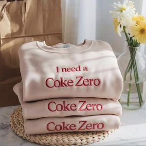 Może przedstawiać: Stos trzech beżowych bluz z napisem "I need a Coke Zero" w kolorze czerwonym. Bluzy są starannie złożone i ułożone na plecionej macie. W tle brązowa papierowa torba i wazon z kwiatami.