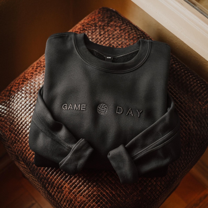 Może przedstawiać: Czarna bluza z napisem "GAME DAY" wyhaftowanym na piersi. Projekt zawiera grafikę siatk&oacute;wki między słowami. Bluza jest złożona na brązowej, teksturowanej powierzchni.
