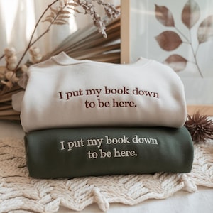Peut inclure: Deux sweats à capuche empilés l'un sur l'autre. Le sweat-shirt du haut est de couleur crème avec le texte "J'ai posé mon livre pour être ici." brodé en brun. Le sweat-shirt du bas est vert foncé avec le même texte brodé en blanc.