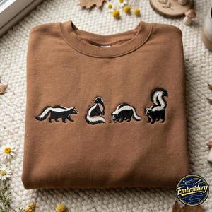 Op de afbeelding: Een bruine sweater met ronde hals voorzien van een borduursel van vier stinkdieren in verschillende houdingen. De stinkdieren zijn afgebeeld met zwart en wit garen en tonen ze lopend, zittend en staand met hun staarten omhoog in een rij.