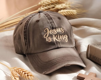 May include: A distressed brown baseball cap with the embroidered text "Jesus is King" in a cursive font. The cap has a curved brim and a worn appearance, suggesting a vintage or casual style. The background includes wheat stalks and a wooden cross.