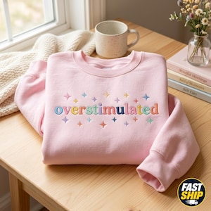 May include: A light pink crewneck sweatshirt featuring the embroidered word "overstimulated" in multicoloured lowercase letters, surrounded by small, colourful embroidered star shapes scattered above and below the text on the front chest area.