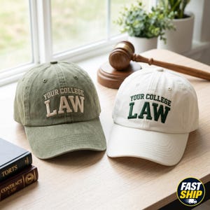 May include: Two baseball caps, one olive green and one white, with the text "YOUR COLLEGE LAW" embroidered on the front. A wooden gavel and books are in the background. The caps are on a light wood surface.