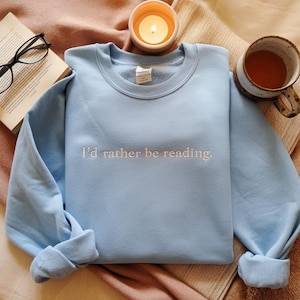 May include: A light blue sweatshirt with the embroidered text "I'd rather be reading." The sweatshirt is displayed with a lit candle, a cup of tea, and a pair of reading glasses next to an open book.