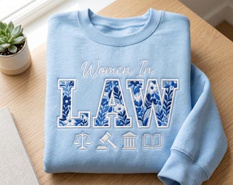 Puede incluir: Sudadera azul claro con el texto "Women In LAW" bordado en blanco. Las letras "LAW" están rellenas con un estampado floral azul y blanco. Debajo del texto hay pequeños iconos blancos de una balanza, martillo, edificio y libro.
