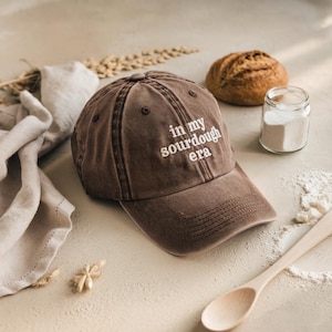 May include: Brown baseball cap with the embroidered text "in my sourdough era". The cap is distressed and has a curved brim. The image also includes a loaf of bread, flour, and a wooden spoon.