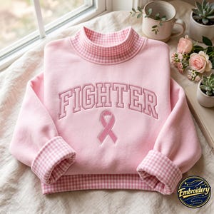 Op de afbeelding: Een lichtroze sweatshirt met een opstaande kraag en boorden in een roze en wit gingham-patroon. De voorzijde toont het woord FIGHTER in gebogen, geborduurd schrift boven een roze bewustzijnslint, alles uitgevoerd in bijpassend roze garen.