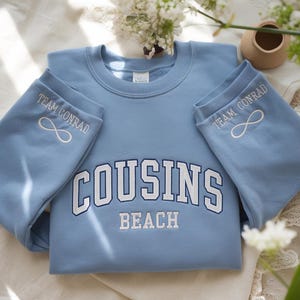 Può includere: Felpa azzurra con la scritta "COUSINS BEACH" ricamata in bianco. Le maniche presentano "TEAM CONRAD" e un simbolo dell'infinito. La felpa è a girocollo.