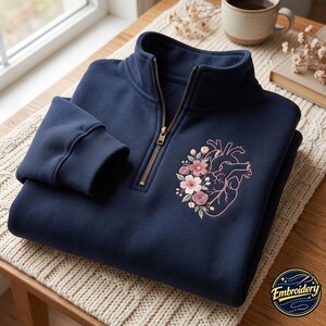 May include: A navy blue quarter-zip sweatshirt featuring an embroidered anatomical heart intertwined with light pink and white flowers on the left chest. The garment has a metal zipper, ribbed cuffs, and a high collar. A circular logo reads Embroidery.