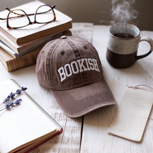Op de afbeelding: Bruine baseballcap met het woord "BOOKISH" in wit geborduurd. De cap ligt op een wit houten oppervlak naast boeken, een koffiekopje en een notitieboek.